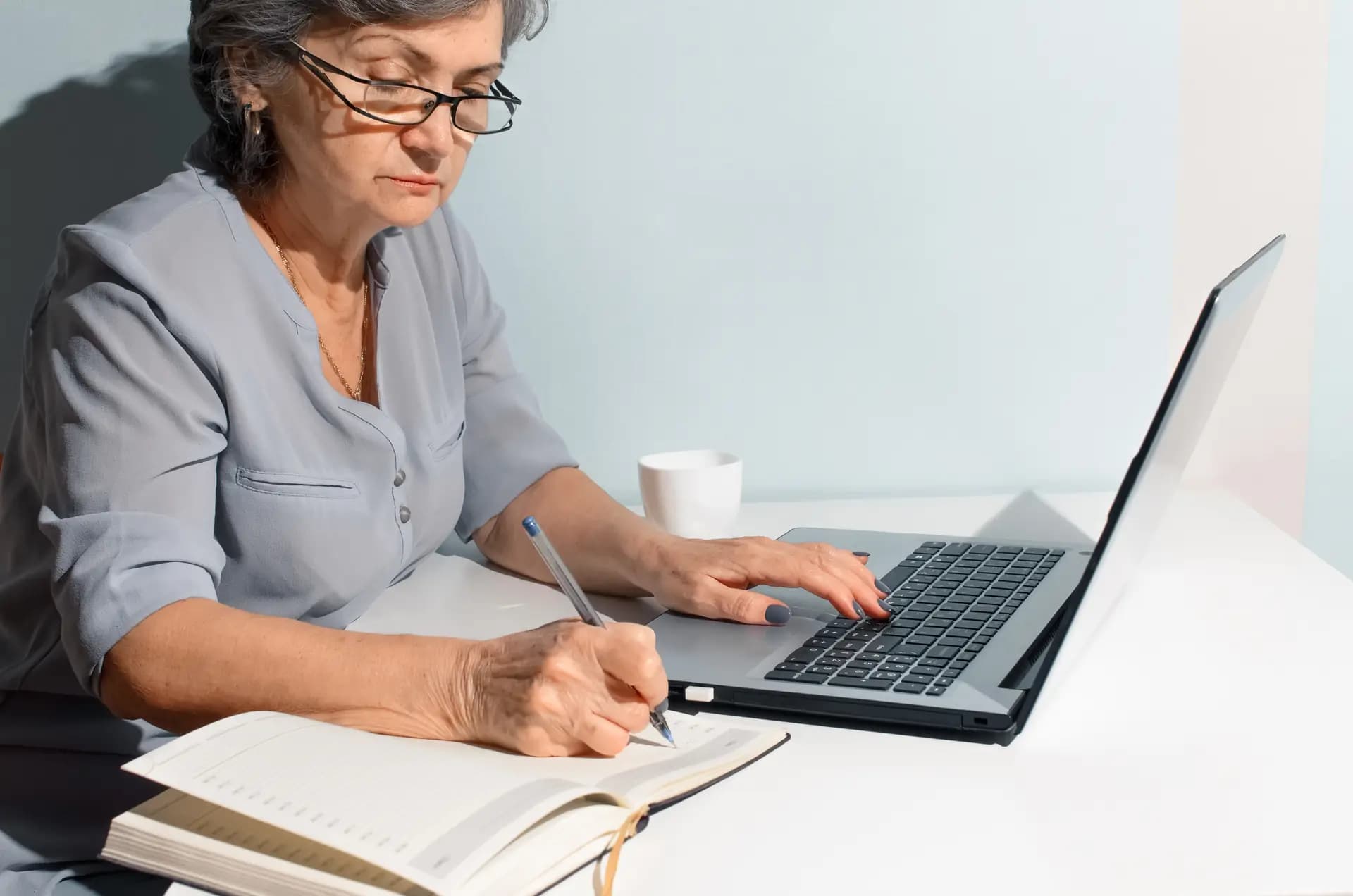 A writer making handwritten notes in a notebook while working at a laptop, with a cup of coffee on a bright desk